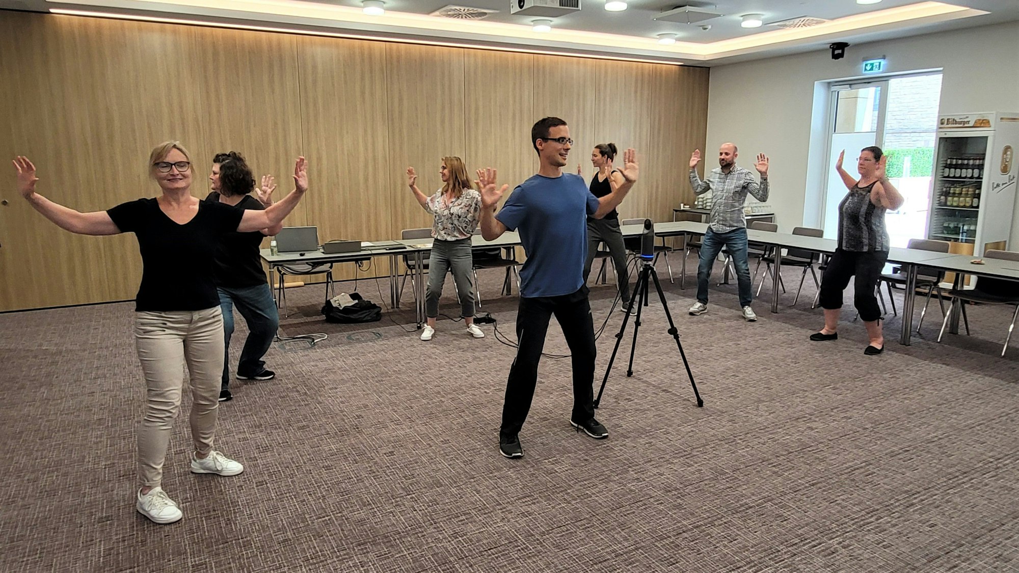 Trainer André Haep (M.) führt mit einer Gruppe Menschen in einem Konferenzraum einen Fitnesskurs durch.