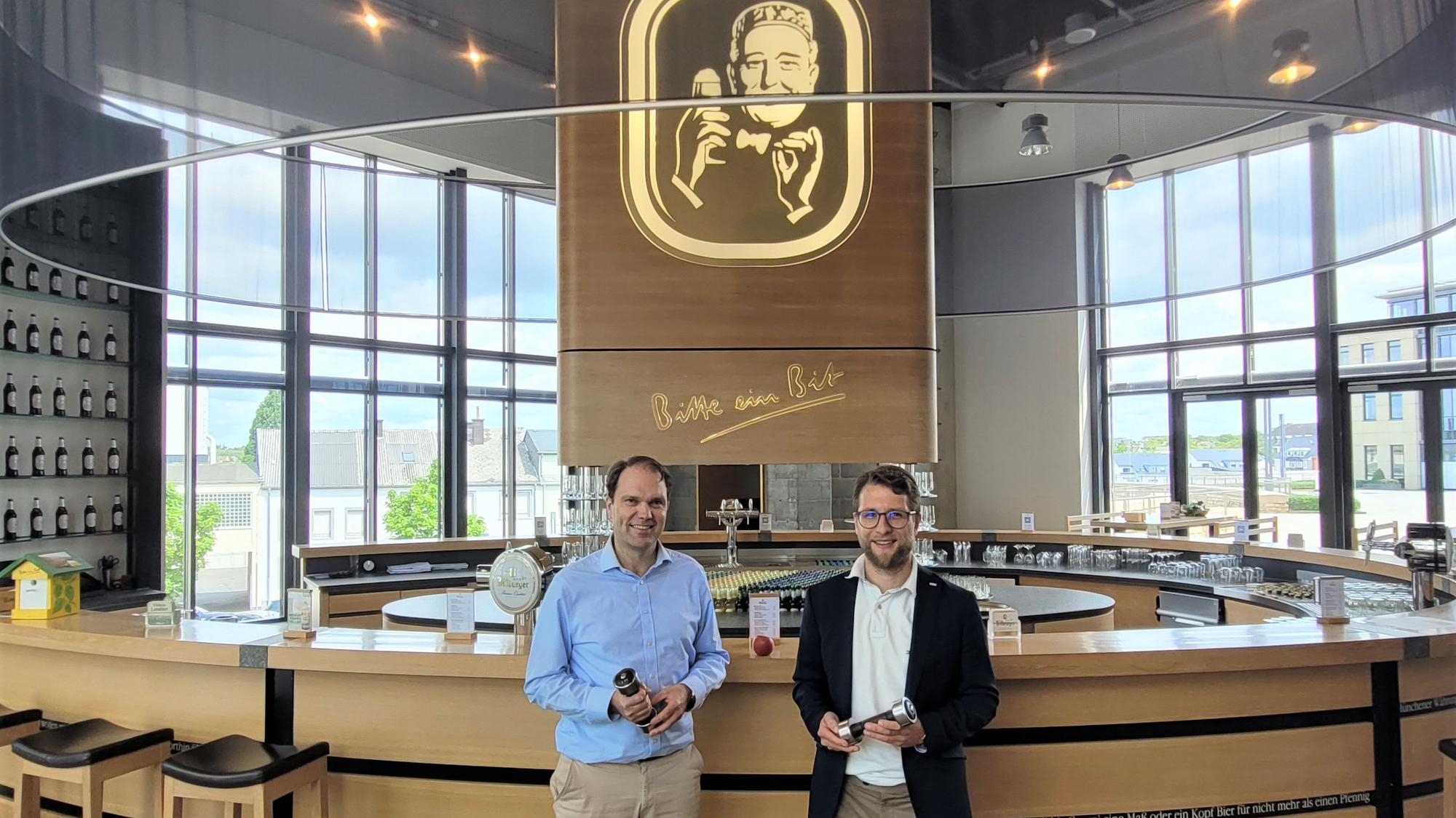 Zwei Männer, Dr. Stefan Schmitz (l.) und Simon Wittig, stehen vor einem Tresen in der Bitburger Brauerei. In der Mitte hängt ein großes Bitburger-Logo.