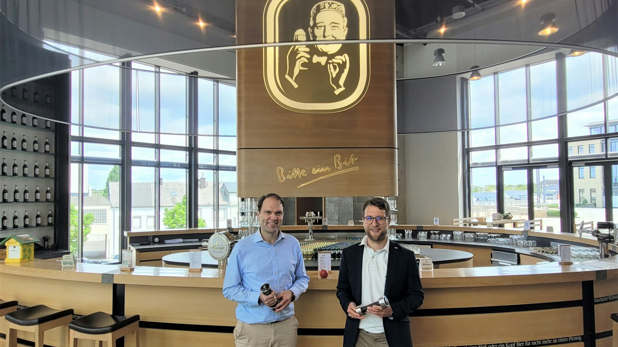 Zwei Männer, Dr. Stefan Schmitz (l.) und Simon Wittig, stehen vor einem Tresen in der Bitburger Brauerei. In der Mitte hängt ein großes Bitburger-Logo.