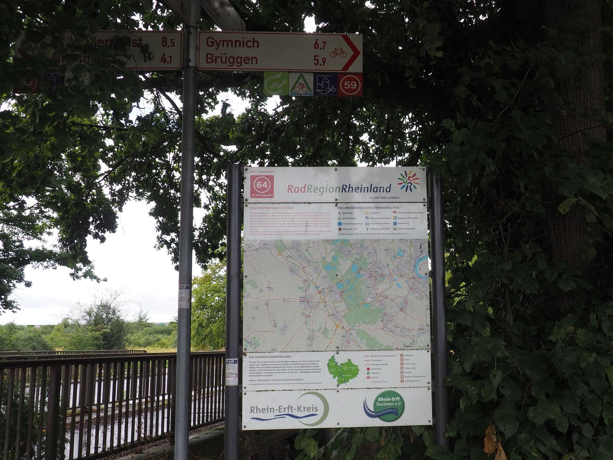 Radweg-Schilder zeigen nach Gymnich und Brüggen, daneben hängt eine Karte.