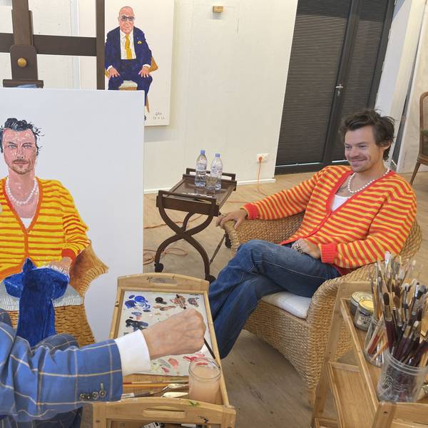 Der berühmte Maler David Hockney (links) malt Harry Styles (rechts). Hockney trägt einen blau karieren Anzug und eine weiße Schiebermütze, Styles eine gelb-rot-gestreifte Strickjacke.