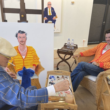 Der berühmte Maler David Hockney (links) malt Harry Styles (rechts). Hockney trägt einen blau karieren Anzug und eine weiße Schiebermütze, Styles eine gelb-rot-gestreifte Strickjacke.