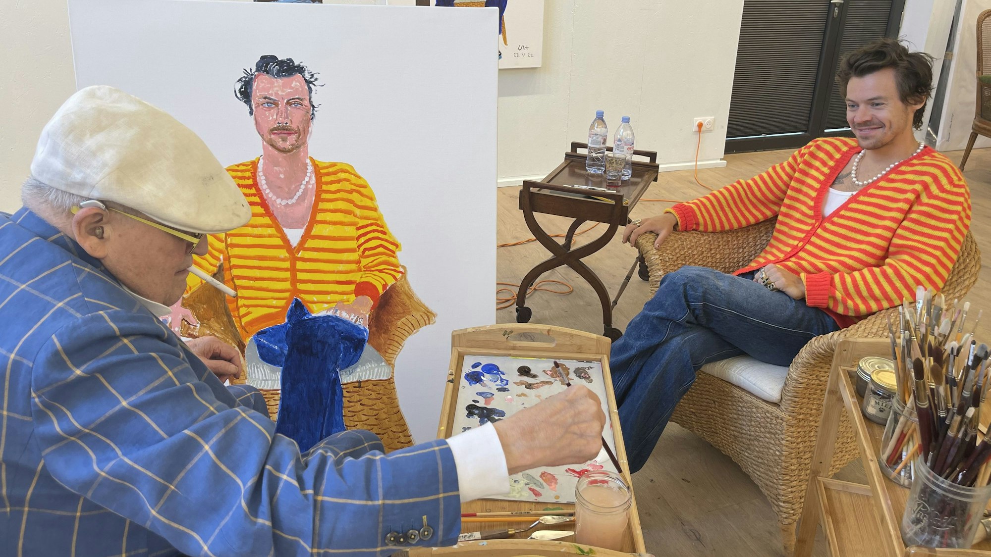 Der berühmte Maler David Hockney (links) malt Harry Styles (rechts). Hockney trägt einen blau karieren Anzug und eine weiße Schiebermütze, Styles eine gelb-rot-gestreifte Strickjacke.