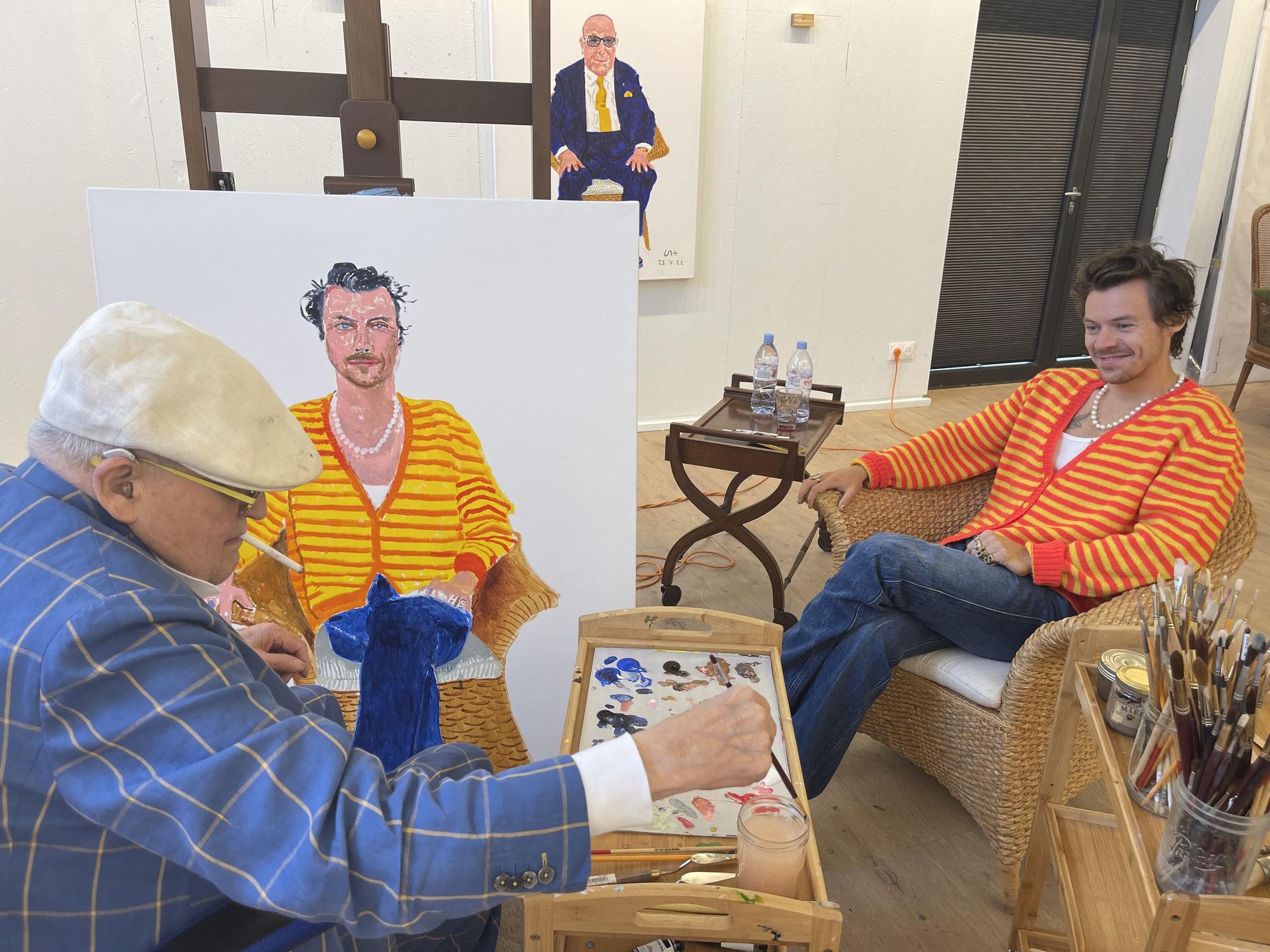 Der berühmte Maler David Hockney (links) malt Harry Styles (rechts). Hockney trägt einen blau karieren Anzug und eine weiße Schiebermütze, Styles eine gelb-rot-gestreifte Strickjacke.