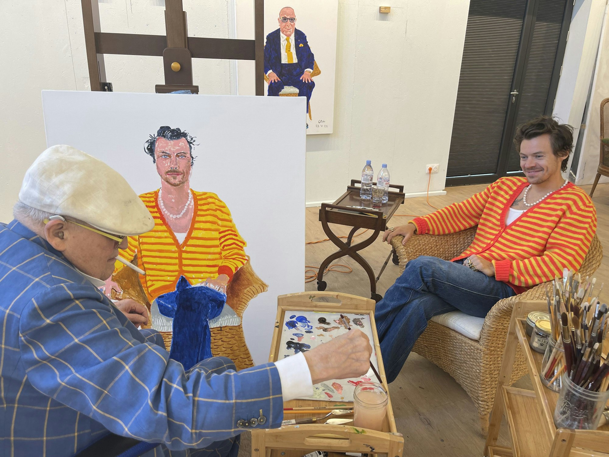 Der berühmte Maler David Hockney (links) malt Harry Styles (rechts). Hockney trägt einen blau karieren Anzug und eine weiße Schiebermütze, Styles eine gelb-rot-gestreifte Strickjacke.