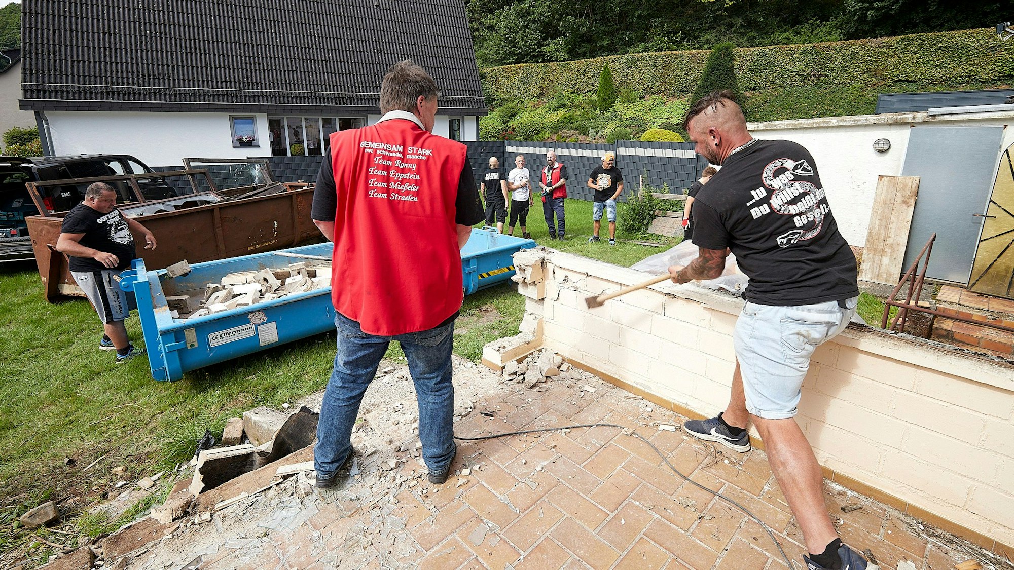 Ein Mann schlägt mit einem Hammer gegen eine niedrige Mauer, die abgerissen wird. Ein weiterer Mann schaut zu.