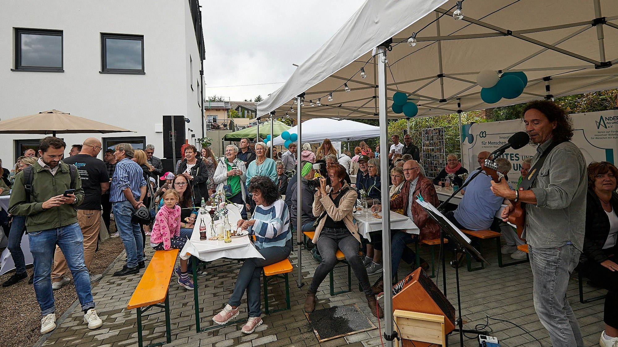 Stephan Brings begleitet sich beim Singen selbst mit der Gitarre. Die Gäste der Einweihungsfeier hören zu, im Hintergrund das neu gebaute Haus