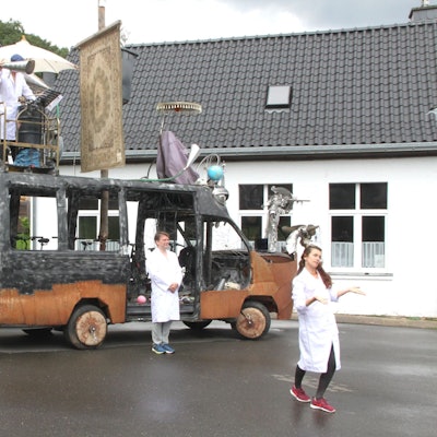 Die futuristische Klima-Galeere des Künstlers Peter Ratz steht auf einem Platz. Zwei Künstler in weißen Kitteln stehen davor, einer darauf.