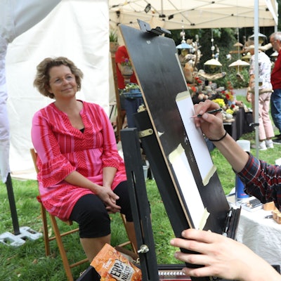 Eine Frau mit Hut zeichnet an einer Staffelei stehend ein Porträt.