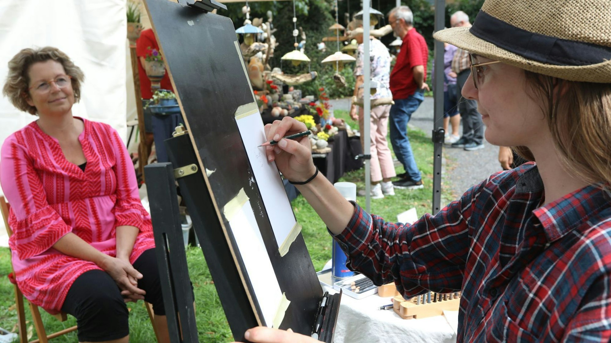 Eine Frau mit Hut zeichnet an einer Staffelei stehend ein Porträt.