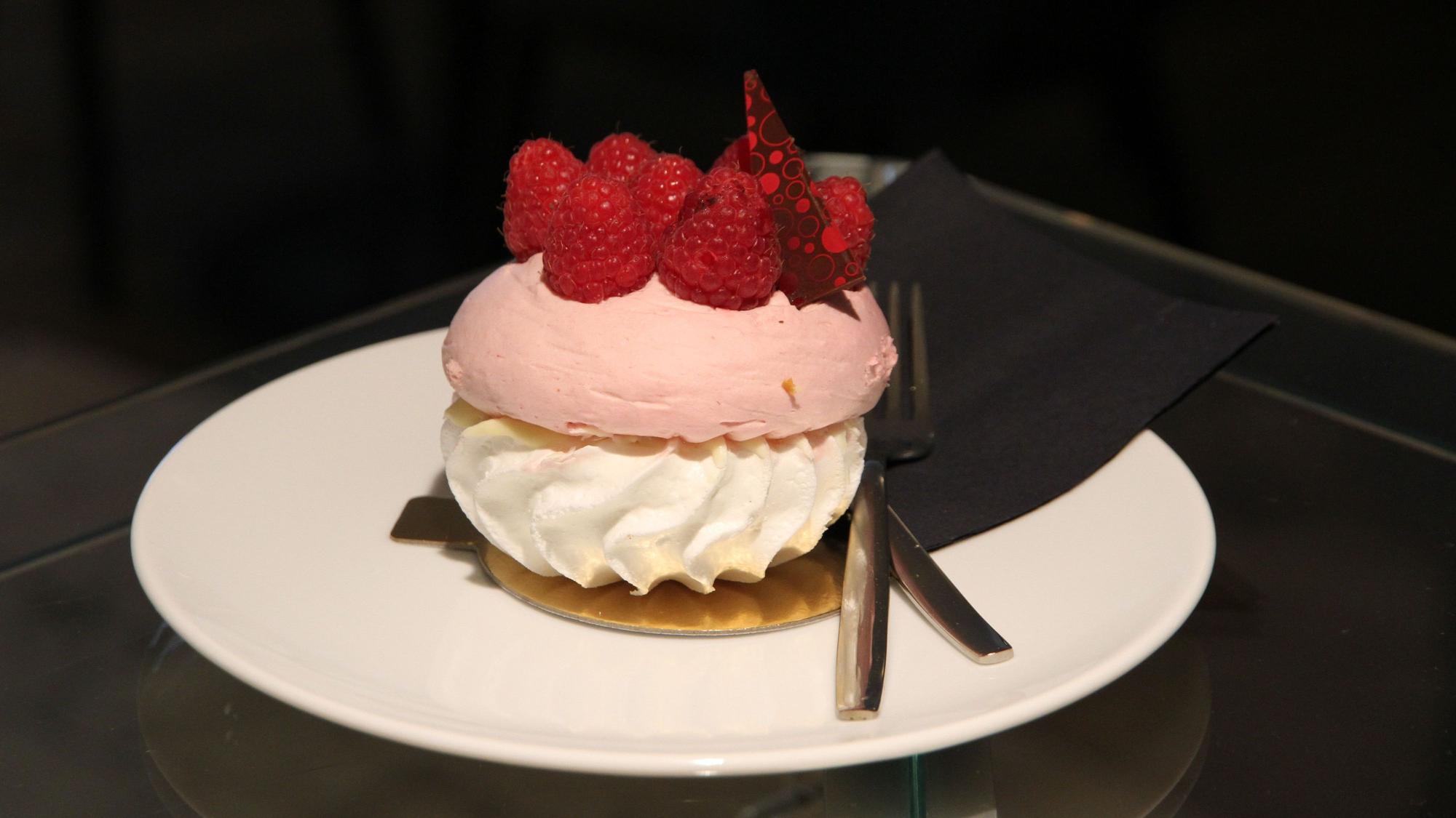 Ein Törtchen aus rosa Baiser, weißer Creme und obenauf Himbeeren auf einem weißen Teller.