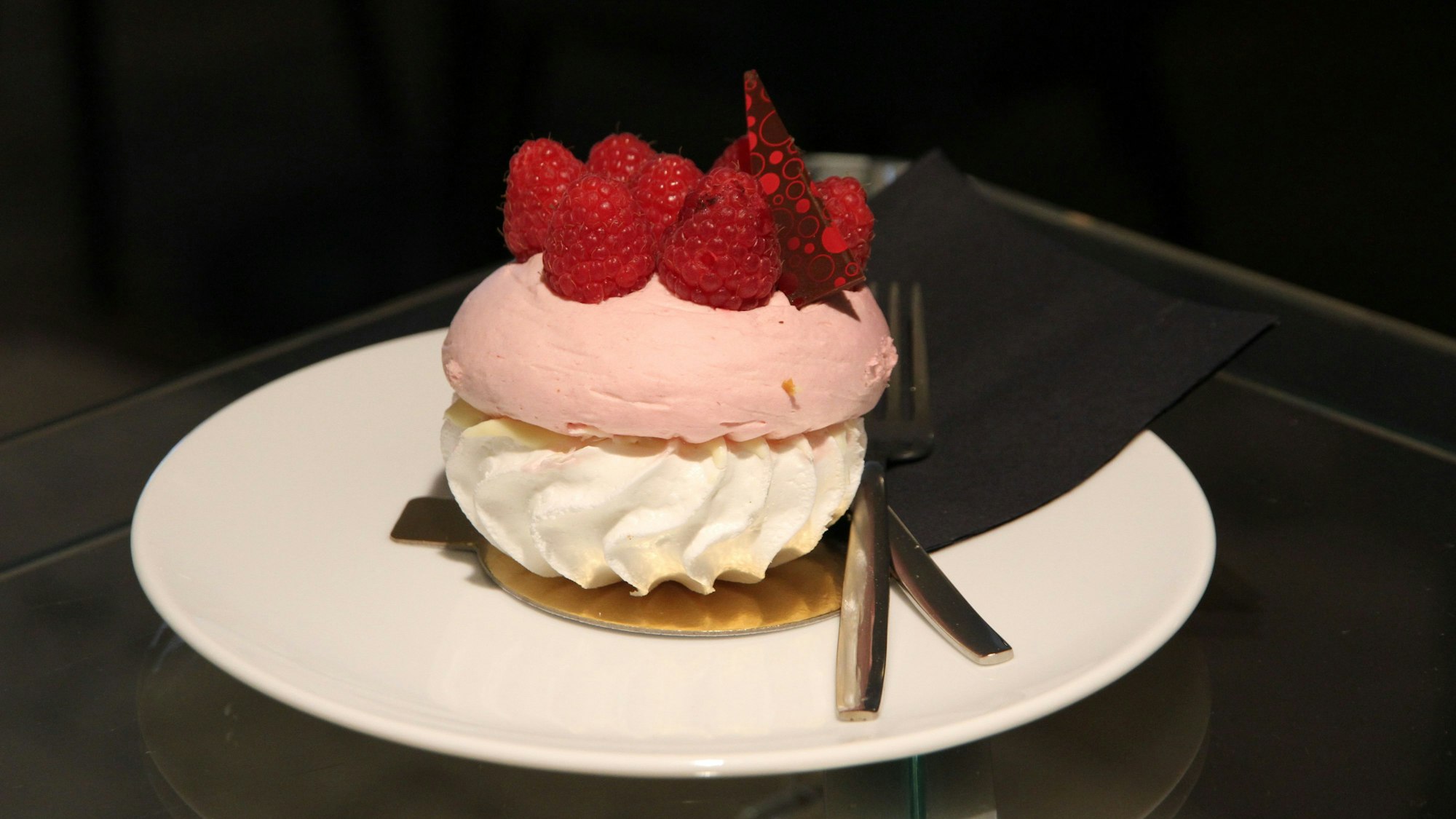 Ein Törtchen aus rosa Baiser, weißer Creme und obenauf Himbeeren auf einem weißen Teller.