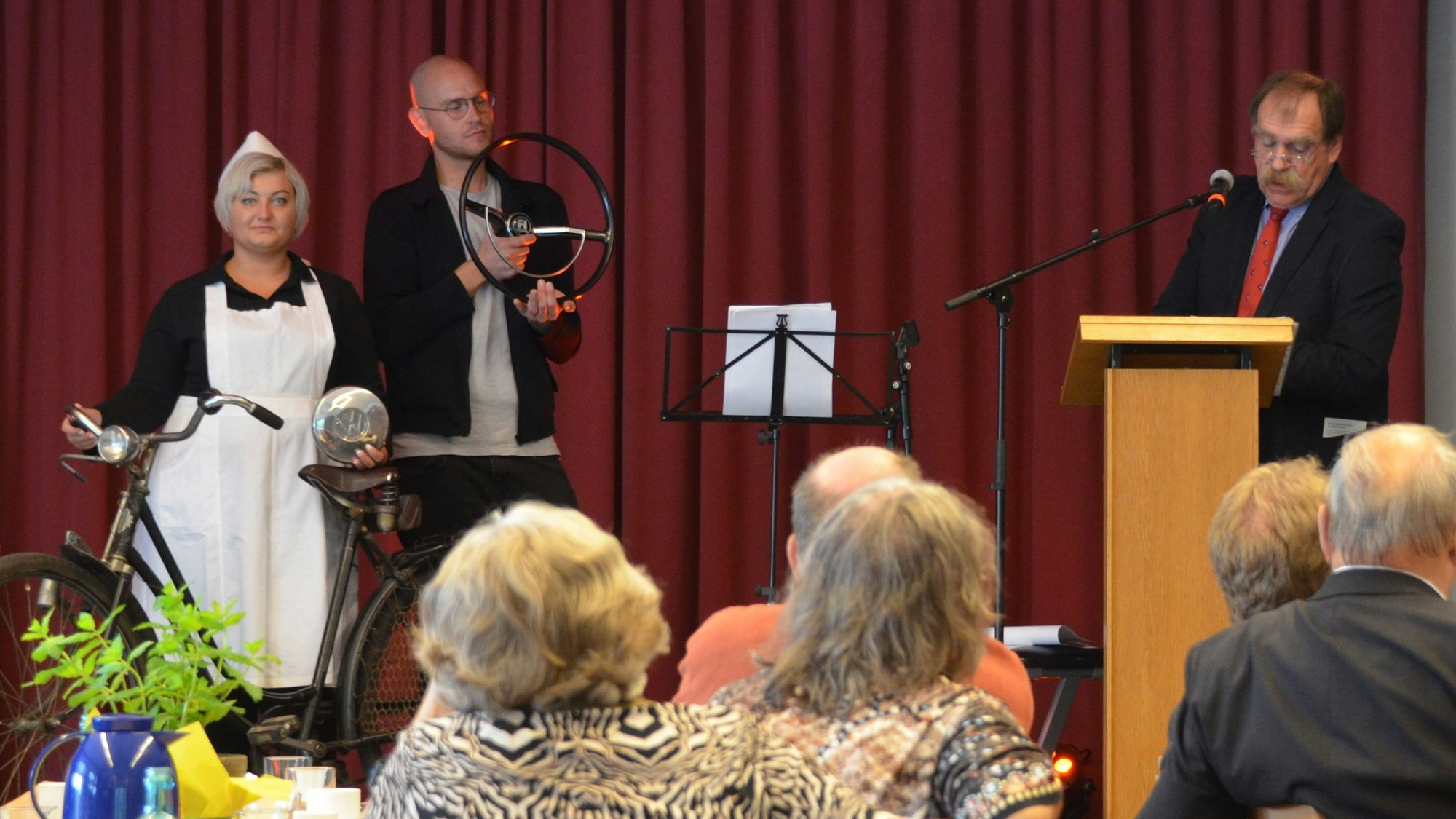 Eine Frau in Schwesterntracht mit Fahrrad und ein Mann mit einem Autolenkrad in der Hand stehen neben dem Vortragenden am Pult.