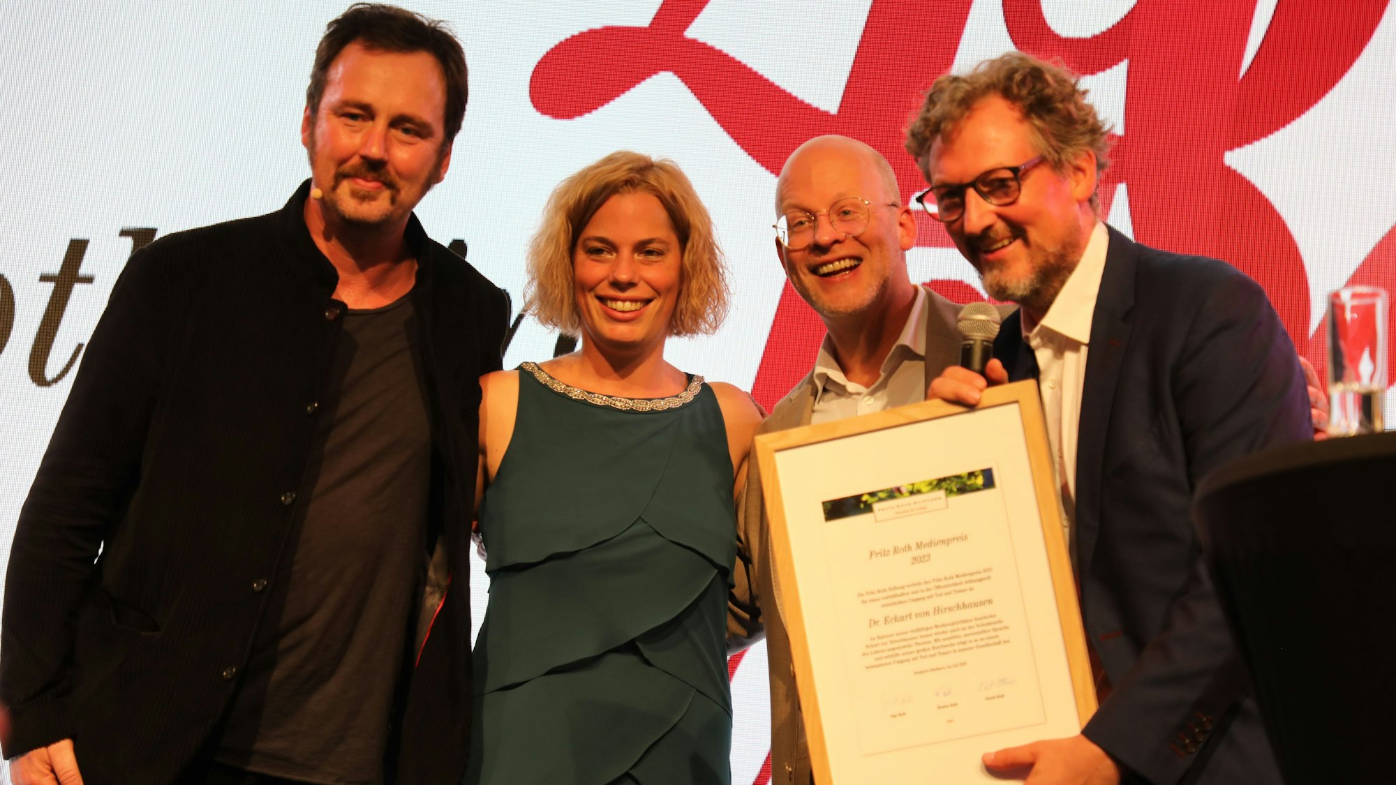 Der Arzt, Fernsehmacher und Kabarettist Dr. Eckhart von Hirschhausen steht mit einer Urkunde in Händen sowie Laudator Eric Wrede, Hanna Roth und Bruder David Roth auf einer Bühne.