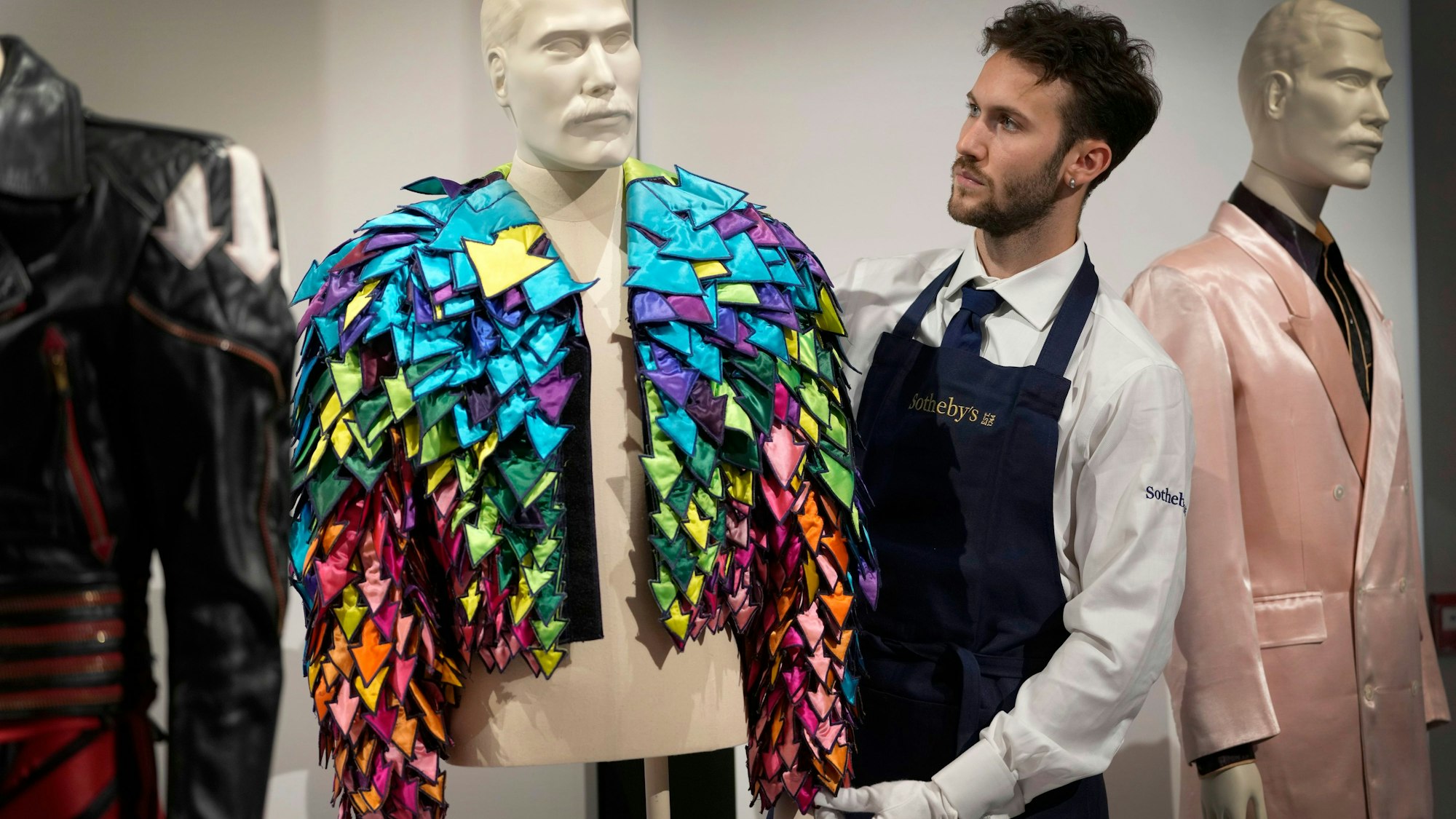 Großbritannien, London: Ein Mitarbeiter von Sotheby's zeigt eine «regenbogenfarbene Jacke mit Pfeil-Applikationen aus Satin von 1982» in den Auktionsräumen von Sotheby's. Mehr als 1.000 persönliche Gegenstände von Freddie Mercury, darunter seine extravaganten Bühnenkostüme, handgeschriebene Entwürfe von «Bohemian Rhapsody» und der Babyflügel, mit dem er die größten Hits von Queen komponierte, werden in einer Ausstellung bei Sotheby's London vor ihrer Versteigerung gezeigt.
