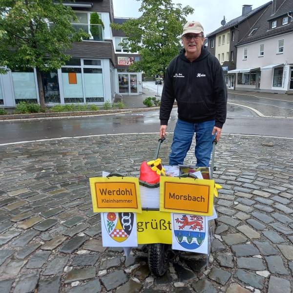 Reinhard Kaldeich steht mit einer mit den Ortsschildern von Werdohl und Morsbach geschmückten Schubkarre auf dem leeren Milly-la-Forêt-Platz in Morsbach.