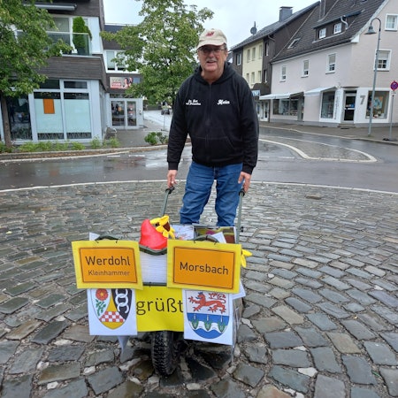 Reinhard Kaldeich steht mit einer mit den Ortsschildern von Werdohl und Morsbach geschmückten Schubkarre auf dem leeren Milly-la-Forêt-Platz in Morsbach.