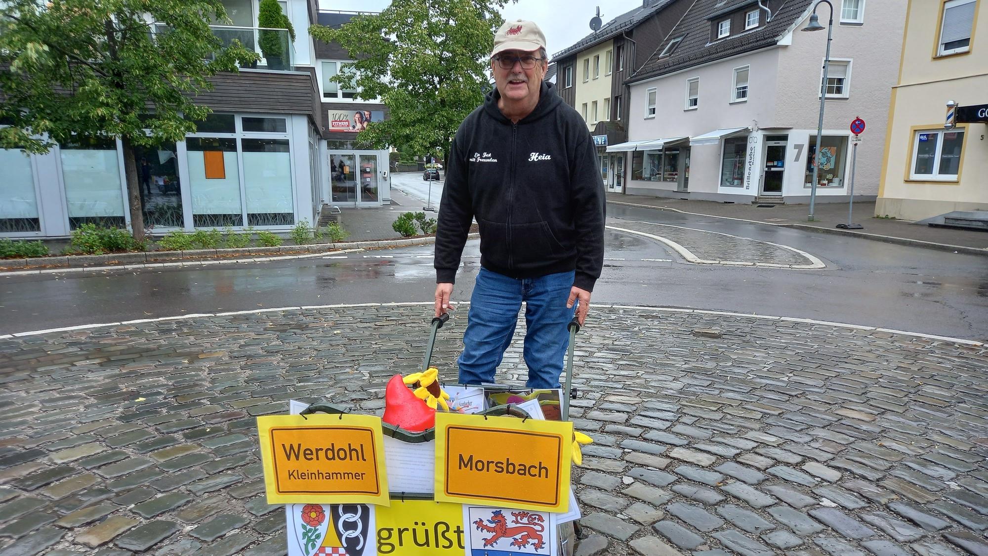 Reinhard Kaldeich steht mit einer mit den Ortsschildern von Werdohl und Morsbach geschmückten Schubkarre auf dem leeren Milly-la-Forêt-Platz in Morsbach.