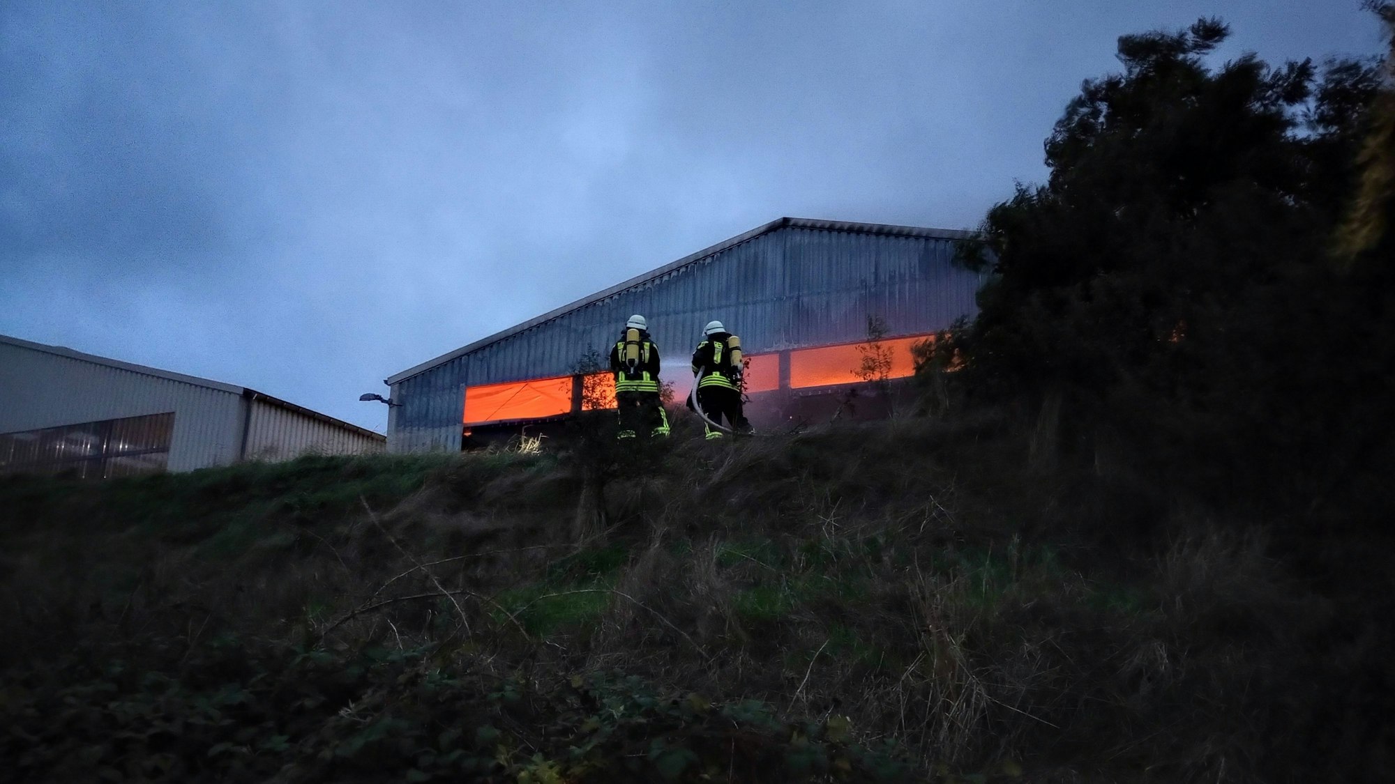Man sieht zwei Feuerwehrleute auf einem Hang, im Hintergrund die Halle, aus der ein Leuchten kommt.