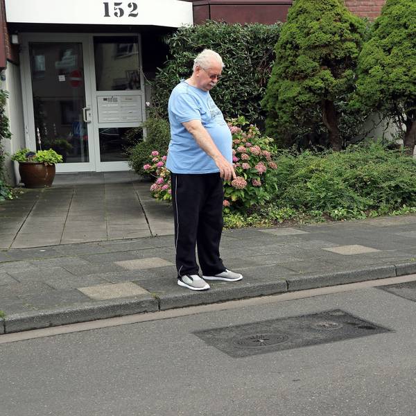 Ein Mann steht vor einem Mehrfamilienhaus an der Straße und zeigt auf geflickten Asphalt.