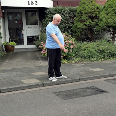 Ein Mann steht vor einem Mehrfamilienhaus an der Straße und zeigt auf geflickten Asphalt.