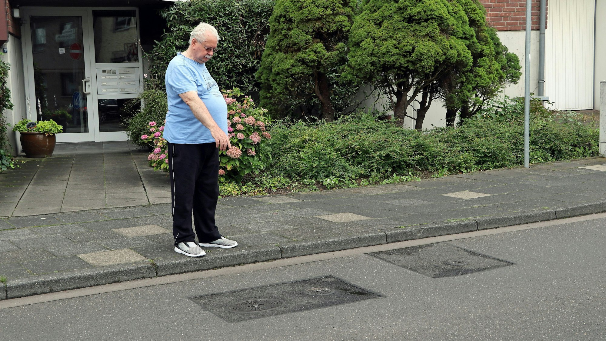 Ein Mann steht vor einem Mehrfamilienhaus an der Straße und zeigt auf geflickten Asphalt.