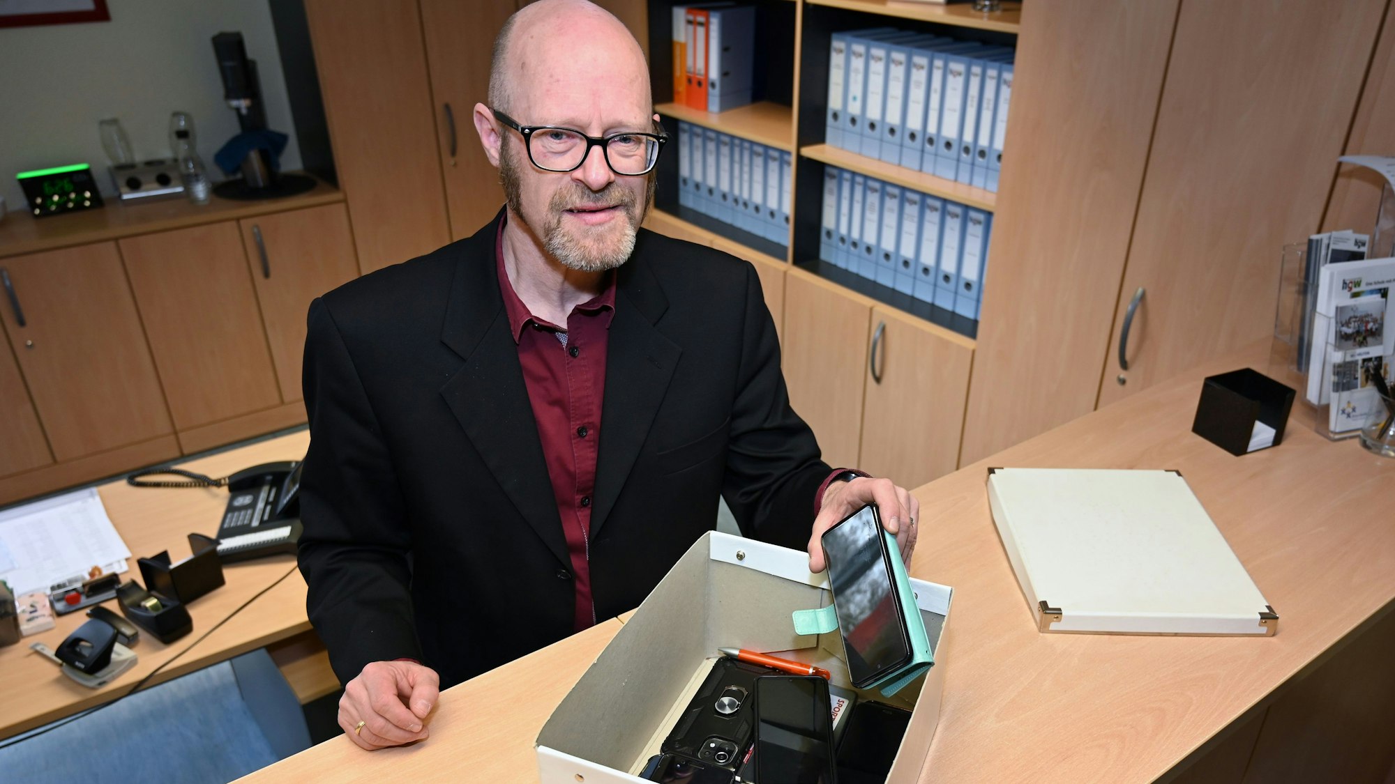 Frank Bohlscheid, Direktor des Hollenberg-Gymnasiums in Waldbröl, steht im Schulsekretariat. Vor ihm auf dem Tresen befindet sich eine Kiste: Dies ist das „Handy-Hotel“, in dem Mobiltelefone von Schülerinnen und Schülern gesammelt werden, die Lehrkräfte zuvor einkassiert haben.