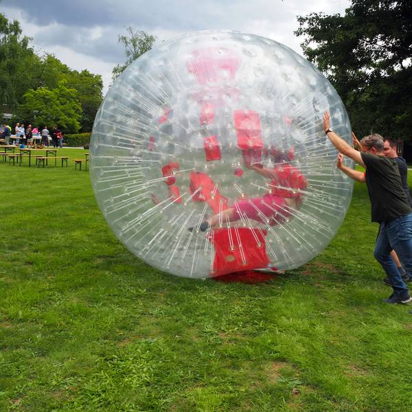 Zwei Männer rollen einen riesigen Plastikball, in dem eine Frau versucht, zu laufen.