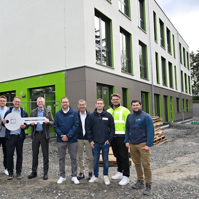 Vor dem Neubau der Modulschule steht der Bürgermeister mit den am Projekt beteiligten Fachleuten. Sie halten einen großen symbolischen Schlüssel in der Hand.