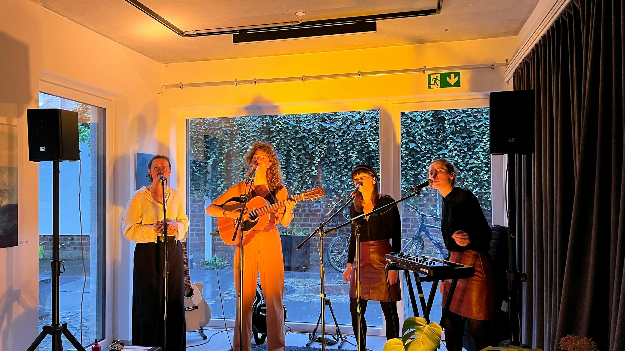 Vier Frauen musizieren vor einer Fensterfront in einem Raum, eine hält eine Gitarre, eine steht am Keyboard, alle vier singen.