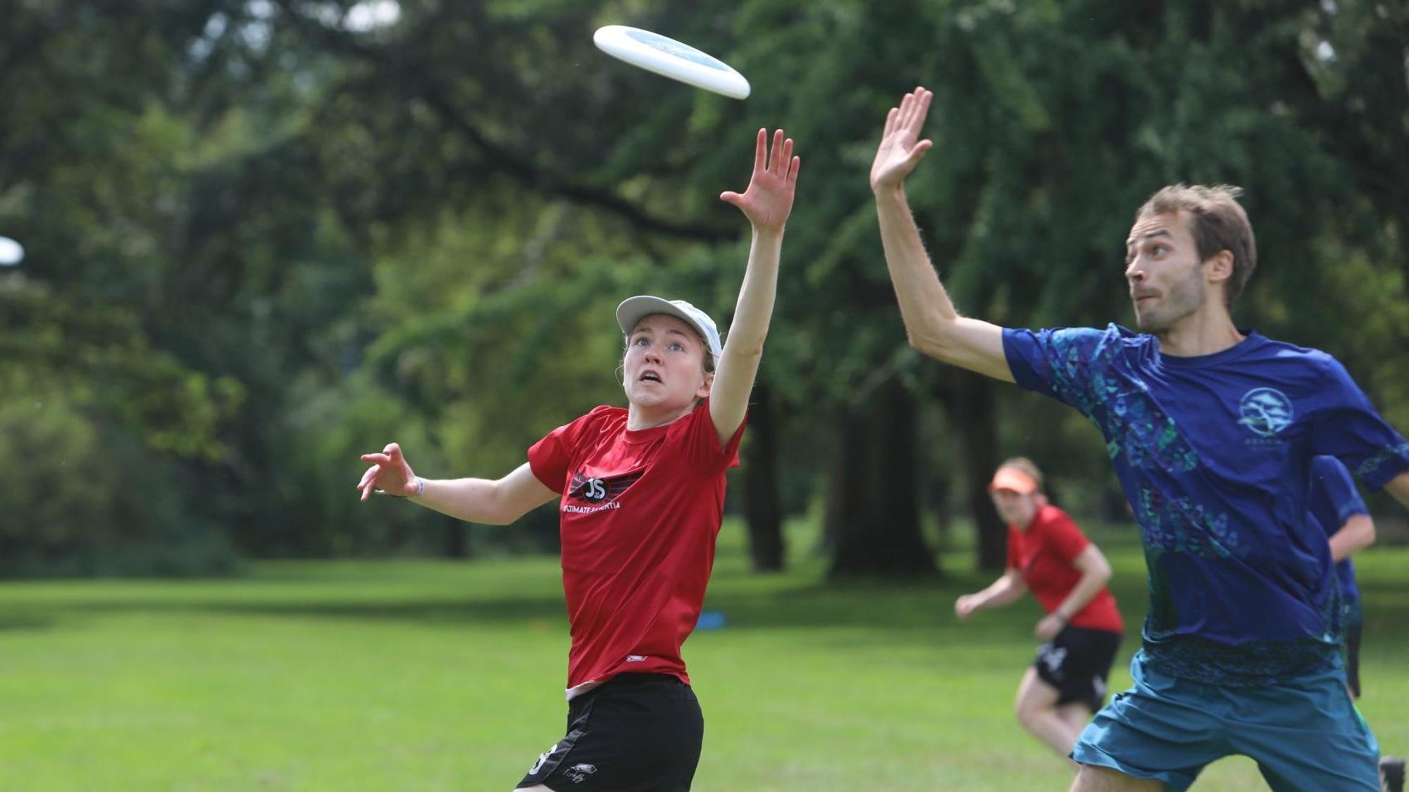 Eine Frau im roten Trikot und ein Mann im blauen Shirt versuchen, eine fliegende Frisbee-Scheibe zu fangen.