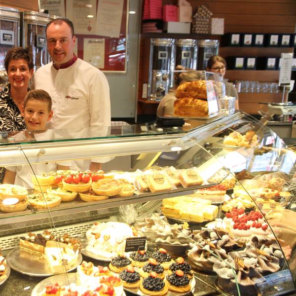 Eine mit Kuchen und Torten gefüllte Kühltheke, dahinter die Eigentümerfamilie und ihr Sohn. Vater und Sohn tragen weiße Konditorenjacken; Im Hintergrund sind alte Kaffeebehälter zu sehen.