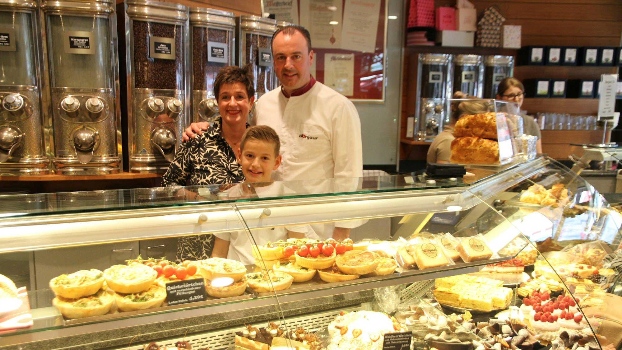 Eine mit Kuchen und Torten gefüllte Kühltheke, dahinter die Eigentümerfamilie und ihr Sohn. Vater und Sohn tragen weiße Konditorenjacken; Im Hintergrund sind alte Kaffeebehälter zu sehen.