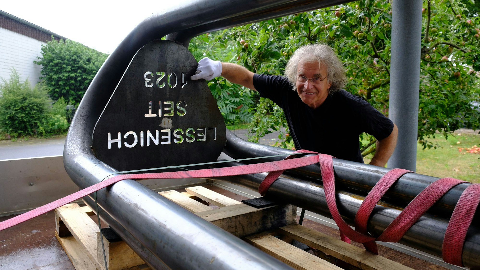 Neben einer Gedenkstele mit der Aufschrift „Lessenich seit 1023“ steht der Künstler Karl Hubert Schmitt. Er trägt einen weißen Handschuh und deutet auf die Inschrift.