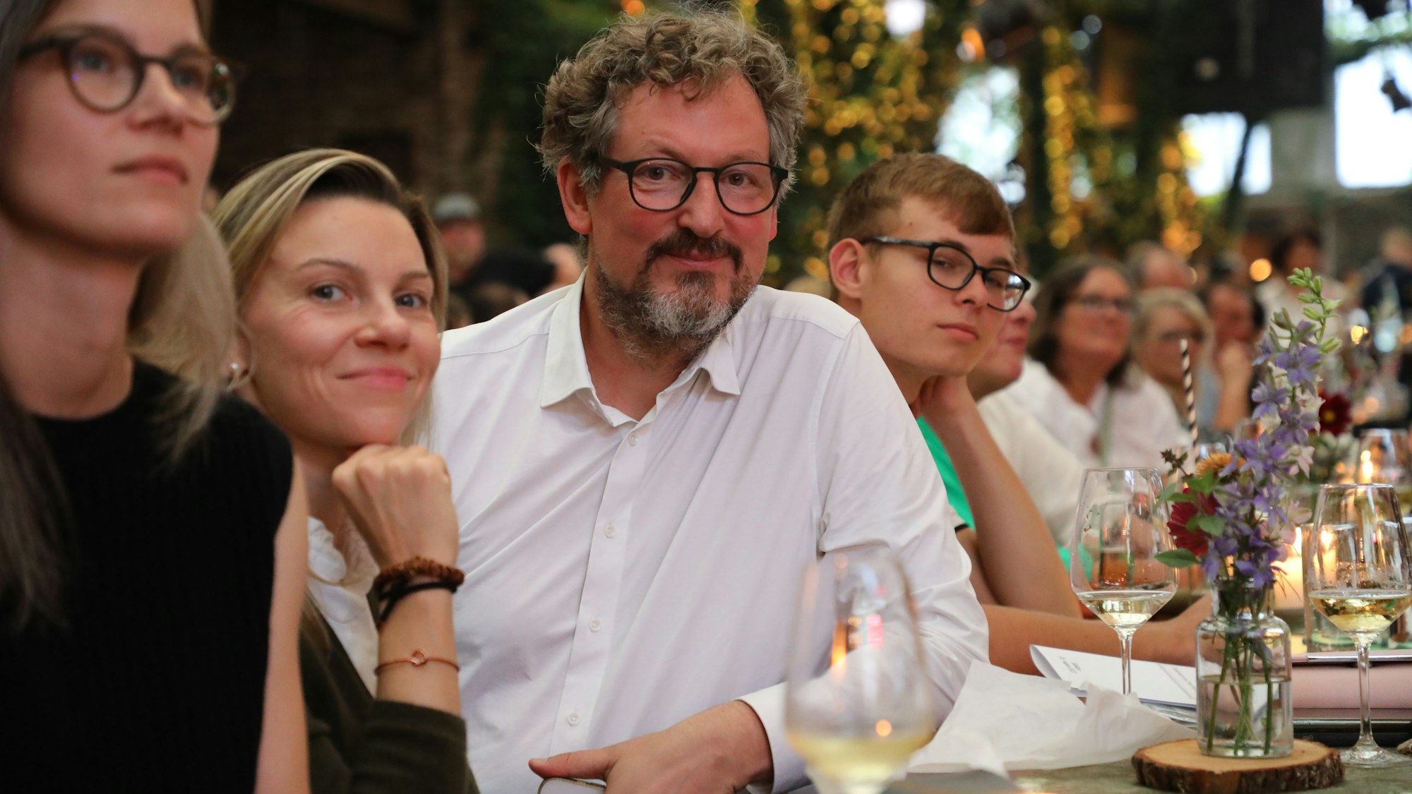 Ein Mann mit Locken, Brille und Bart sitzt im Publikum an einem Tisch und schaut in die Kamera.