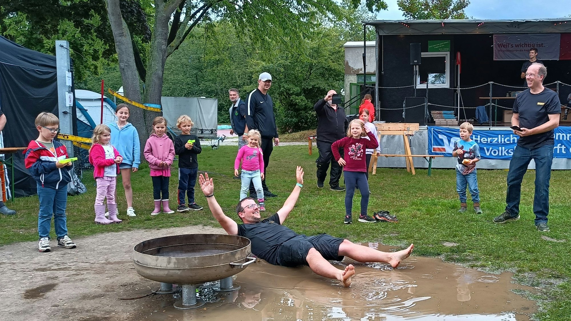 Das Bild zeigt einen Vater, der sich in eine Pfütze wirft. Kinder stehen um ihn herum.