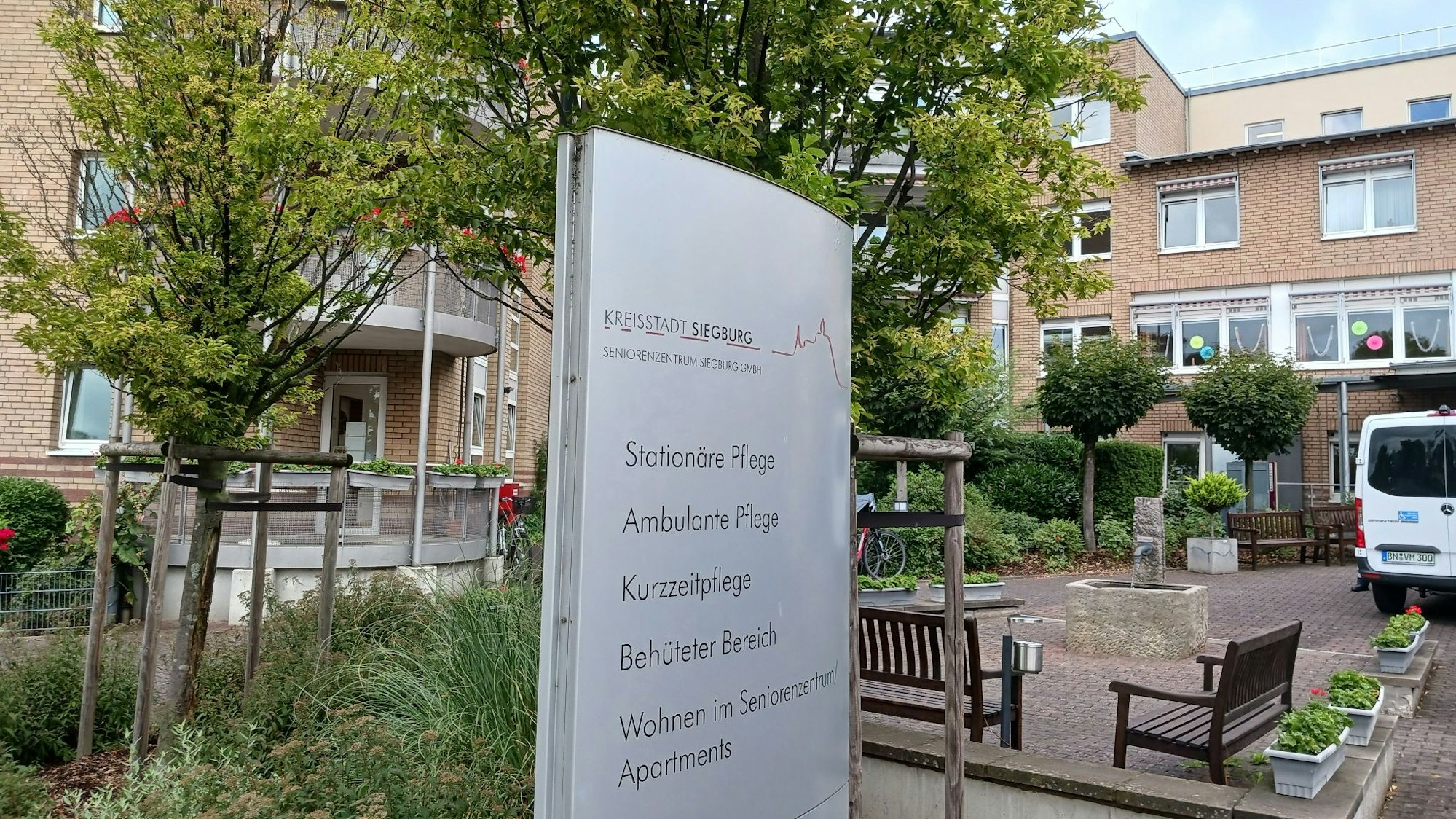 Ein großes Schild vor dem Seniorenzentrum weist auf die unterschiedlichen Abteilungen hin. Im Hintergrund stehen Bänke und Bäume.