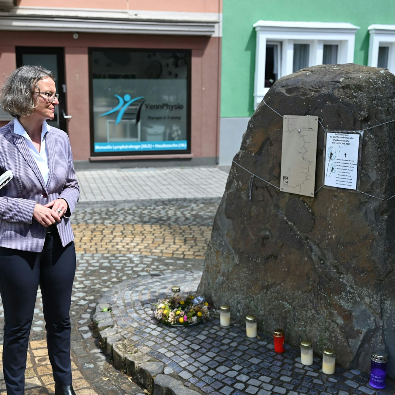 Ina Scharrenbach blickt auf den Gedenkstein. Daran hängt eine Liste mit den Namen der in der Stadt Schleiden bei der Flut ums Leben gekommenen Menschen. Am Fuß des Steins brennen Kerzen.