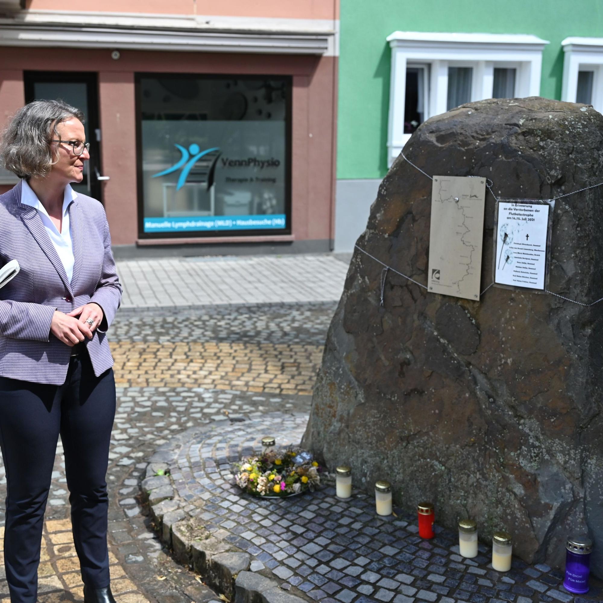 Ina Scharrenbach blickt auf den Gedenkstein. Daran hängt eine Liste mit den Namen der in der Stadt Schleiden bei der Flut ums Leben gekommenen Menschen. Am Fuß des Steins brennen Kerzen.