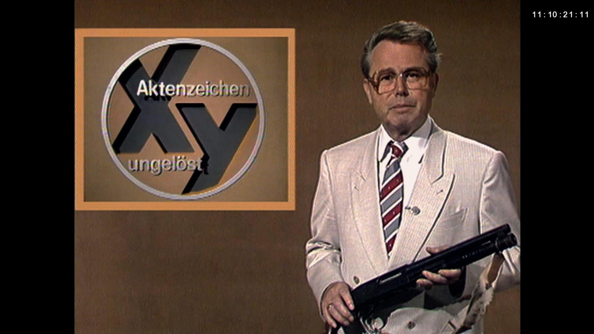 Eduard Zimmermann vor beigem Hintergrund, mit einer Waffe in der Hand im Studio von „Aktenzeichen XY ... ungelöst“.