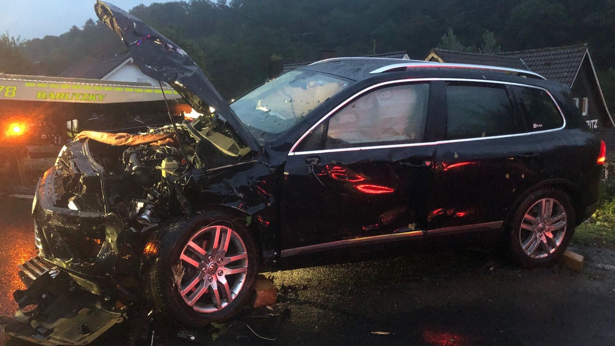Ein schwer beschädigtes Auto steht auf einer Straße in der Dämmerung, im Hintergrund steht ein Abschleppfahrzeug.
