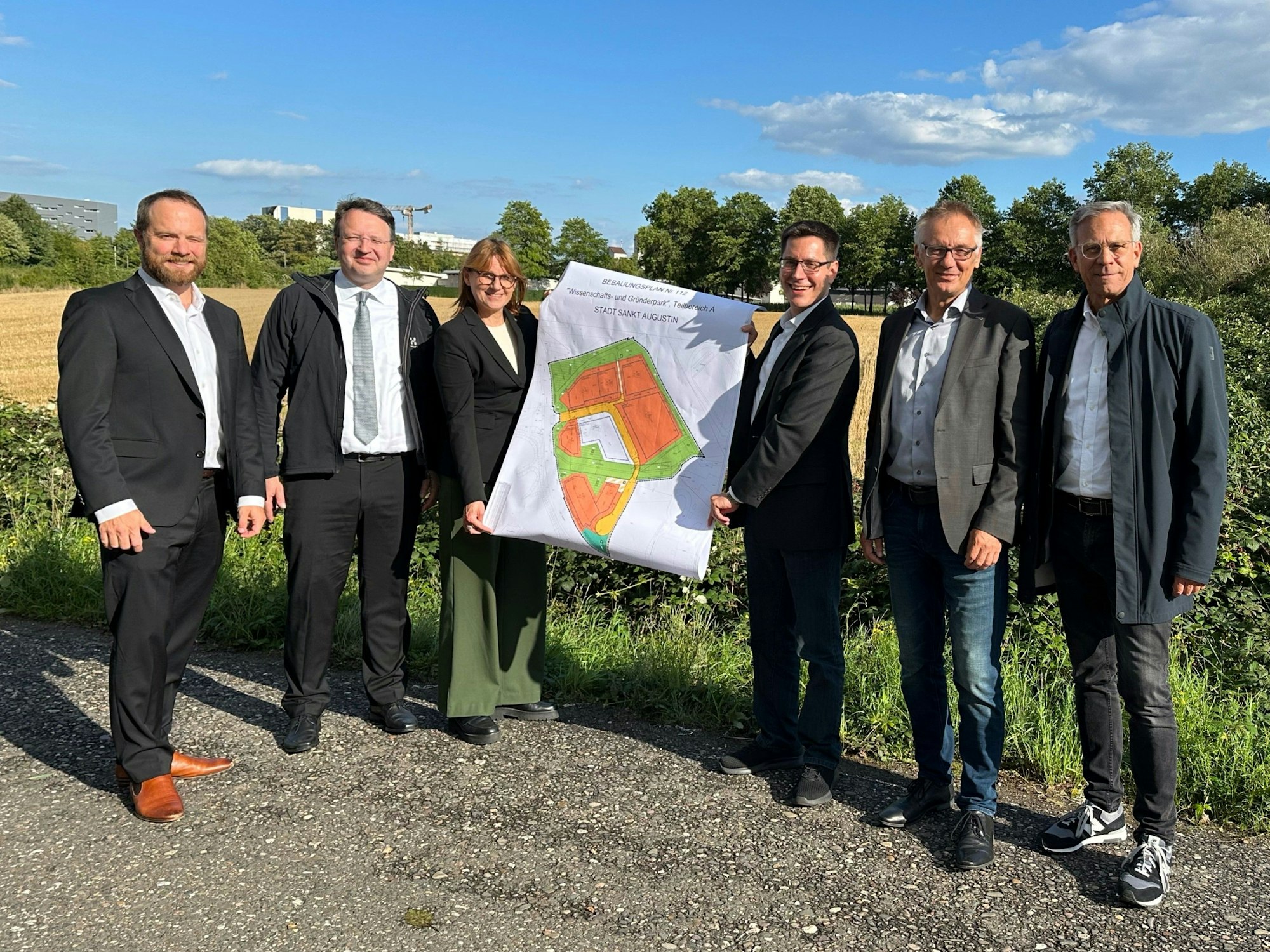 Sechs Menschen halten einen Plan mit einer Zeichnung des Geländes in die Höhe.