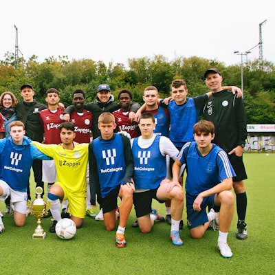 Die Finalisten des von der „Rheinflanke“ organisierten „Nippes-Veedelscup 2023", die „Ukraine Pittbulls“ und „Hope Rheinflanke“ stehen im Nippeser Olympia Stadion, vor ihnen auf dem Boden der Pokal zu Ehren des verstorbenen Fabian S.