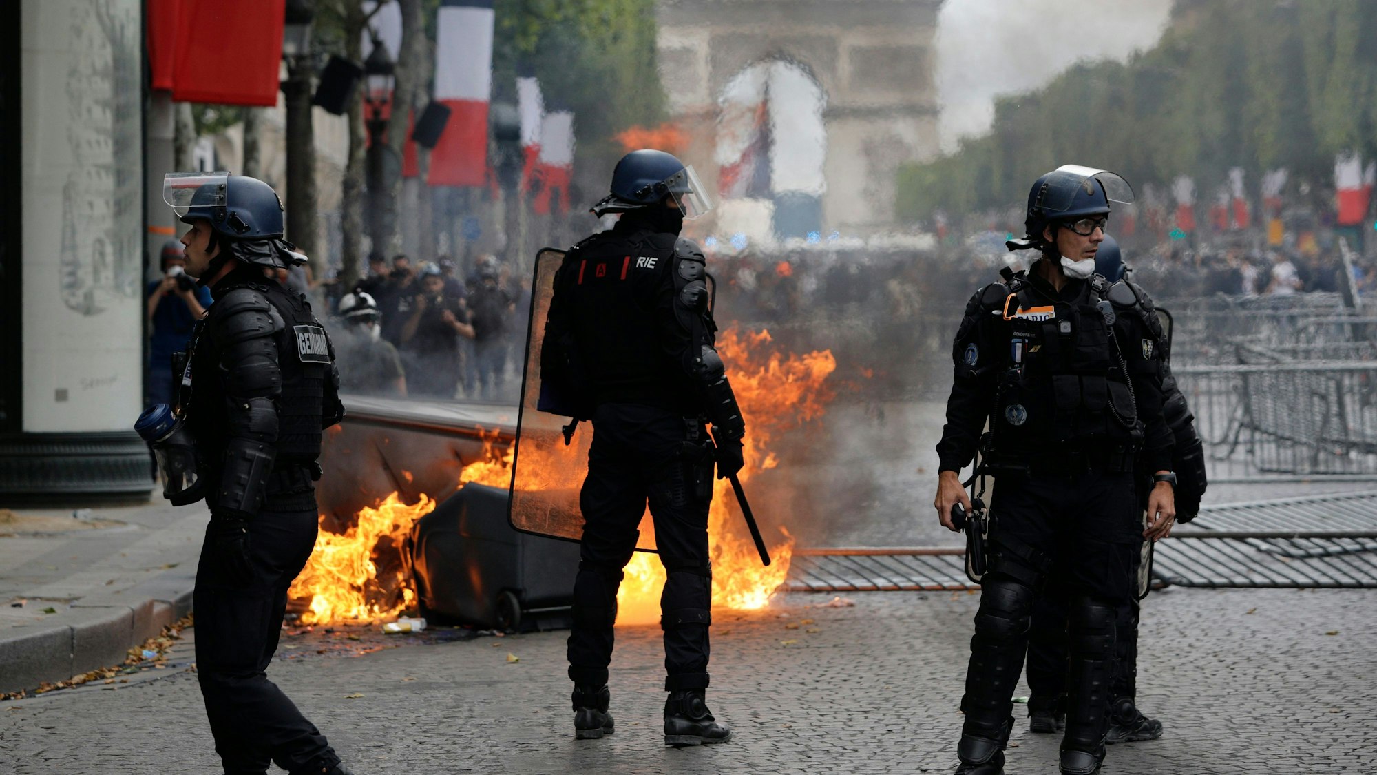 Bereitschaftspolizisten stehen vor einem brennenden Müllcontainer auf der Prachtstraße Champs-Élysées während einer Demonstration am Rande der Pariser Militärparade zum Nationalfeiertag.
