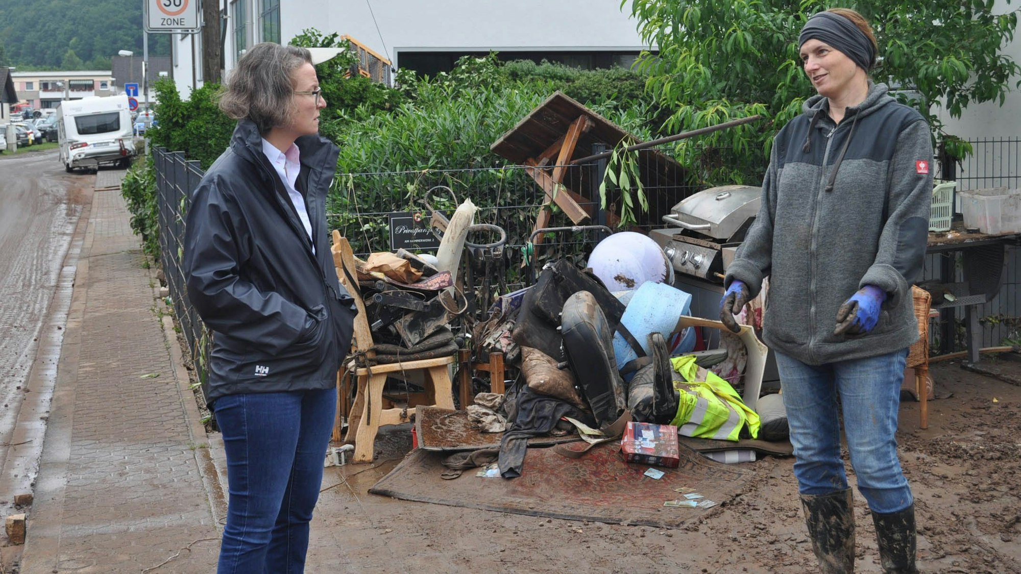 Ministerin Ina Scharrenbach im Gespräch mit einer Betroffenen in Kall. Im Hof liegen Schlamm und zerstörter Hausrat.