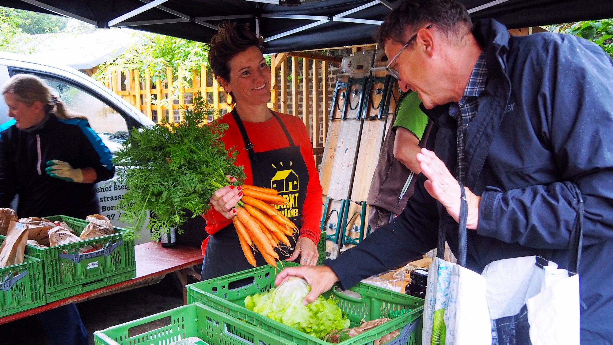 Ein Mann steht an einem Marktstand und prüft Gemüse. Die Verkäuferin hält einen Bund Möheren..