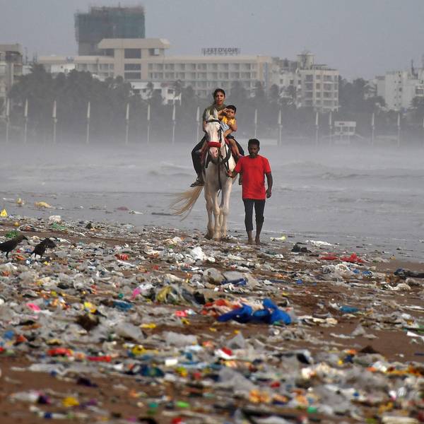 Ein Mann führt ein Pferd mit einer Frau, die ihr Kind im Arm hält, entlang des Strands von Mumbai. Der Strand ist mit Müll, vor allem Kunststoffe, übersät.