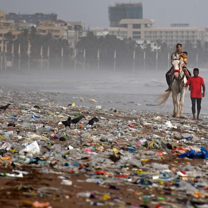 Ein Mann führt ein Pferd mit einer Frau, die ihr Kind im Arm hält, entlang des Strands von Mumbai. Der Strand ist mit Müll, vor allem Kunststoffe, übersät.