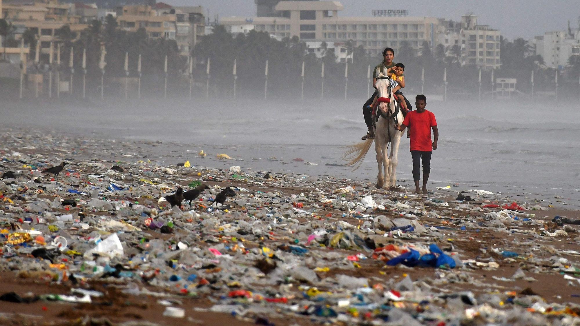 Ein Mann führt ein Pferd mit einer Frau, die ihr Kind im Arm hält, entlang des Strands von Mumbai. Der Strand ist mit Müll, vor allem Kunststoffe, übersät.