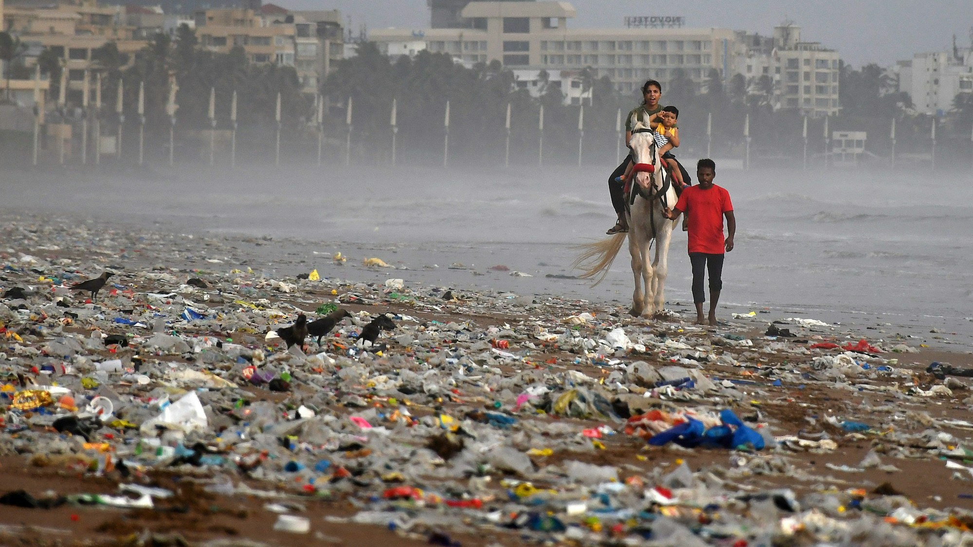 Ein Mann führt ein Pferd mit einer Frau, die ihr Kind im Arm hält, entlang des Strands von Mumbai. Der Strand ist mit Müll, vor allem Kunststoffe, übersät.